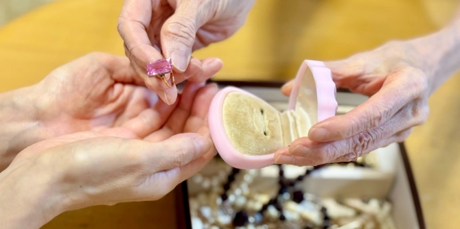 Close-up of two hands handling a pink gemstone ring over an open pink ring box with a cushioned interior.